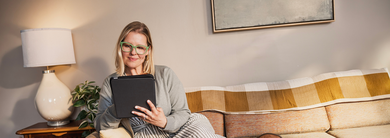 Elderly man with gray hair and glasses, smiling while using a tablet in a cozy room. He wears a blue plaid shirt. A terrarium is beside him.