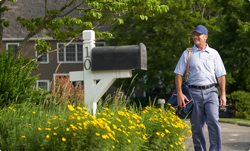 Smiling USPS letter carrier walking up to a mailbox carrying a mailbag.