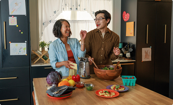 A couple laughing while preparing a salad together in a kitchen.